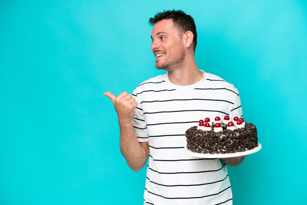 Young caucasian man holding birthday cake isolated on blue background pointing to the side to present a product