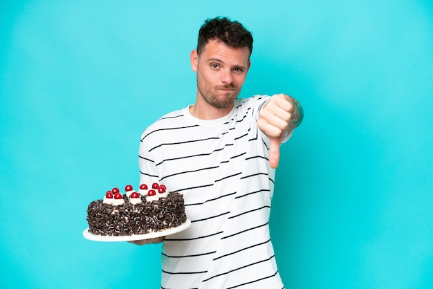 Young caucasian man holding birthday cake isolated on blue background showing thumb down with negative expression