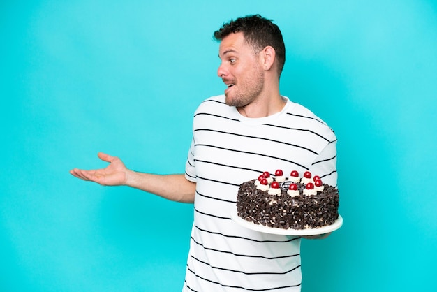 Giovane uomo caucasico che tiene la torta di compleanno isolata su sfondo blu con espressione sorpresa mentre guarda il lato