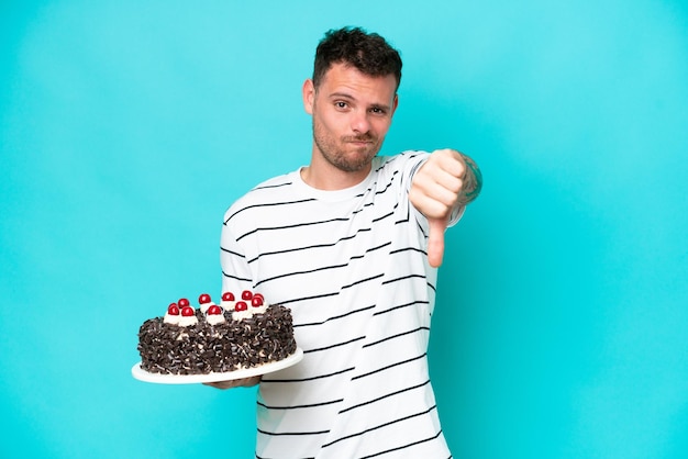 사진 부정적인 표정으로 엄지 손가락을 보여주는 파란색 배경에 고립 된 생일 케이크를 들고 젊은 백인 남자