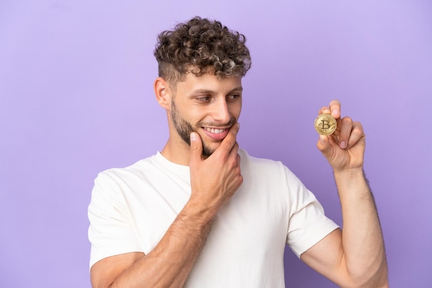 Young caucasian man holding a Bitcoin isolated on purple background looking to the side and smiling