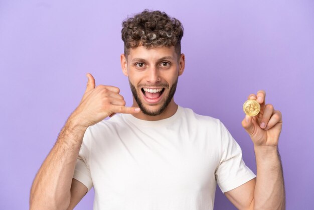 Young caucasian man holding a Bitcoin isolated on purple background making phone gesture Call me back sign