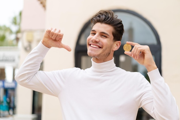 Young caucasian man holding a Bitcoin at outdoors proud and selfsatisfied