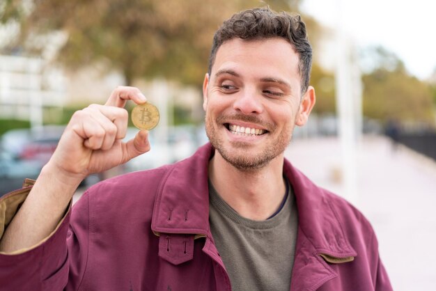 Young caucasian man holding a Bitcoin at outdoors with happy expression