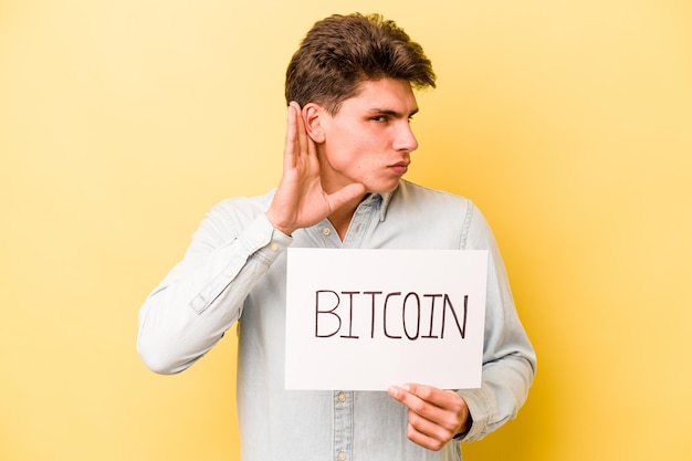 Young caucasian man holding bitcoin placard isolated on yellow background trying to listening a gossip