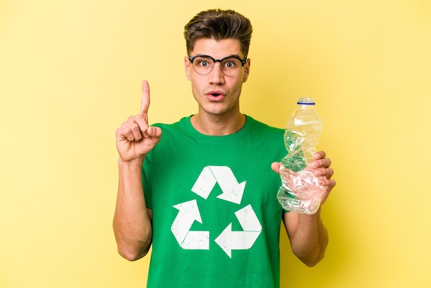 いくつかの素晴らしいアイデア、創造性の概念を持っている黄色の背景に分離されたリサイクルするプラスチックのボトルを保持している若い白人男性。