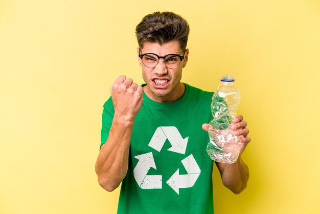 Young caucasian man holding a bottle of plastic to recycle isolated on yellow background showing fist to camera, aggressive facial expression.