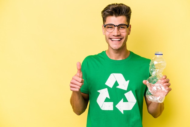Молодой кавказский мужчина держит пластиковую бутылку для переработки, изолированную на желтом фоне, улыбаясь и поднимая большой палец вверх