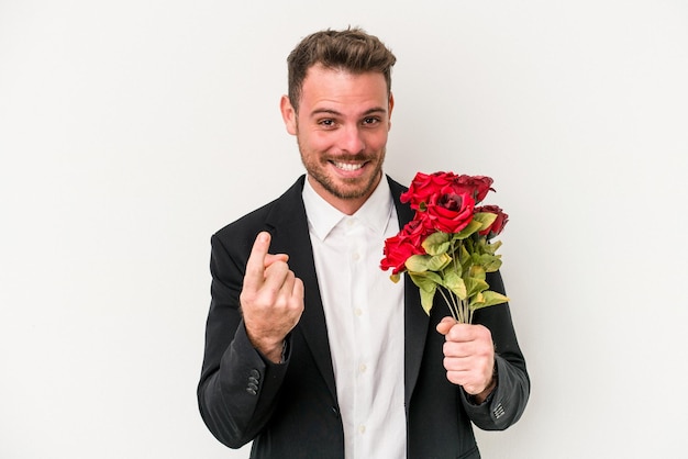 Young caucasian man holding bouquet of flowers isolated on white background pointing with finger at you as if inviting come closer.