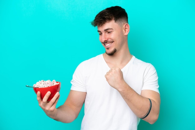 Young caucasian man holding a bowl of cereals isolated on blue background celebrating a victory