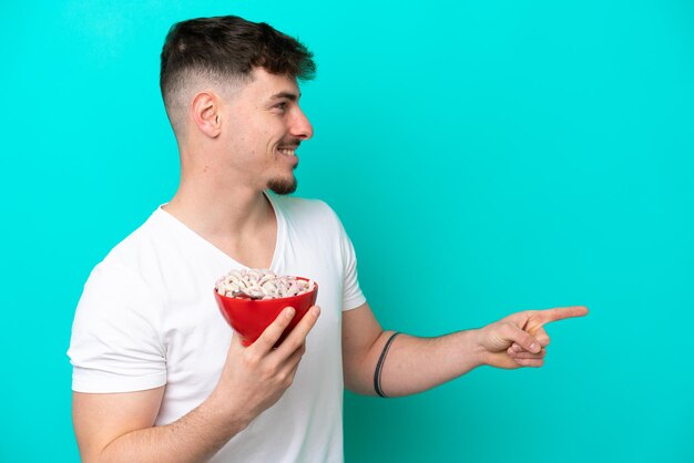 Young caucasian man holding a bowl of cereals isolated on blue\
background pointing to the side to present a product