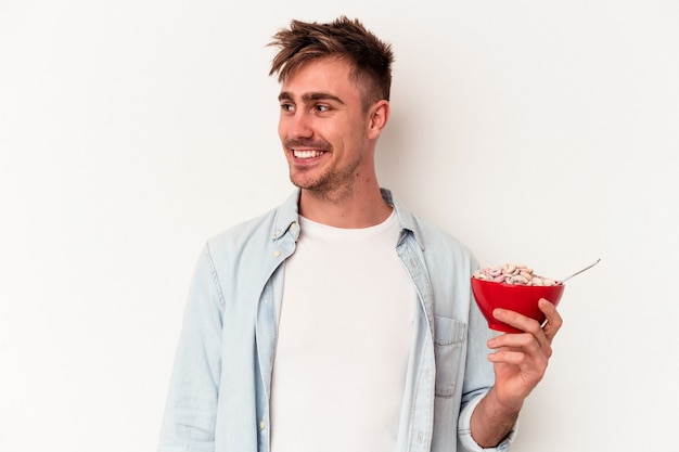 Young caucasian man holding a bowl of cereals isolated on white background looks aside smiling, cheerful and pleasant.