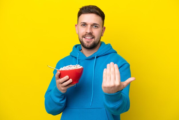 Young caucasian man holding a bowl of cereals isolated on yellow background inviting to come with hand Happy that you came