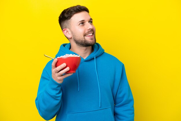 Young caucasian man holding a bowl of cereals isolated on yellow background looking up while smiling
