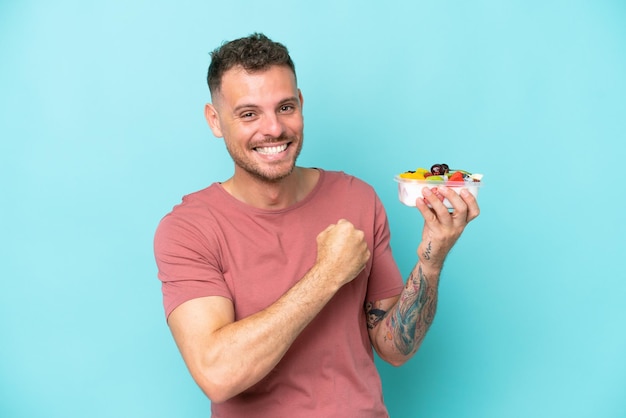 Young caucasian man holding a bowl of fruit isolated on blue background celebrating a victory