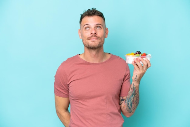 Young caucasian man holding a bowl of fruit isolated on blue background and looking up