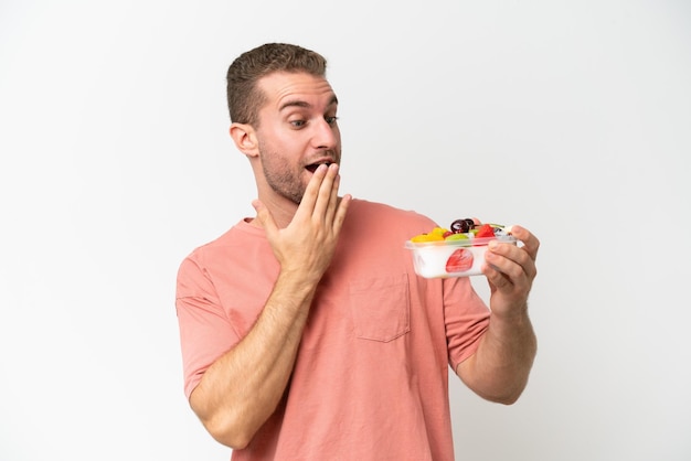 Young caucasian man holding a bowl of fruit isolated on white background with surprise and shocked facial expression