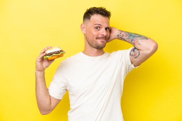 Young caucasian man holding a burger isolated on yellow background having doubts