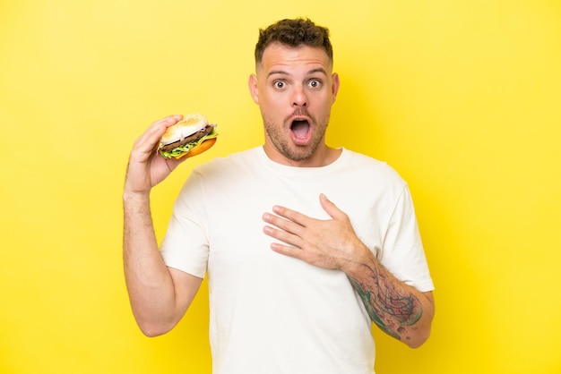 Молодой кавказец, держащий бургер на желтом фоне, удивлен и шокирован, глядя вправо