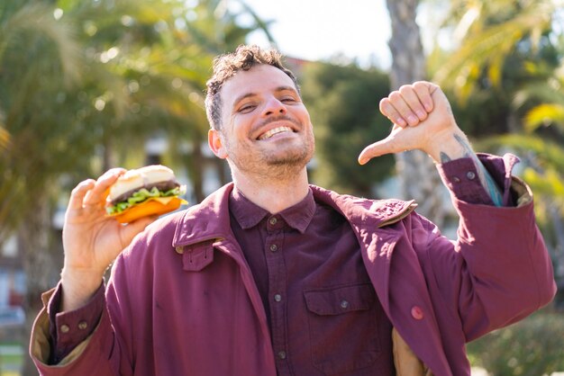 Photo young caucasian man holding a burger at outdoors proud and selfsatisfied