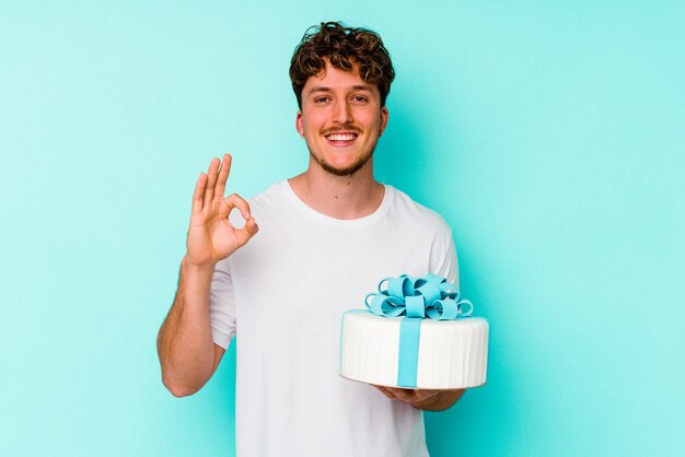 Young caucasian man holding a cake isolated on blue background cheerful and confident showing ok gesture.