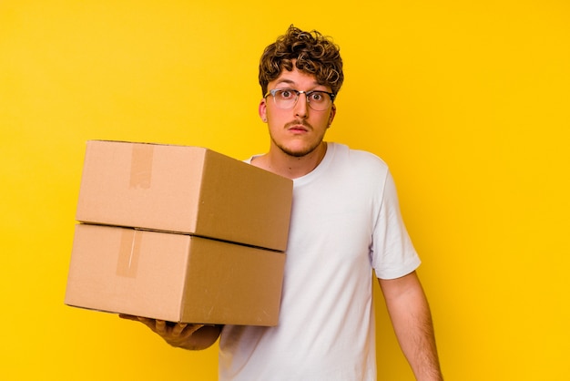 Young caucasian man holding a cardboard box isolated on yellow background shrugs shoulders and open eyes confused.