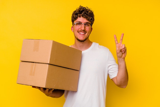 Young caucasian man holding a cardboard box isolated on yellow wall showing number two with fingers.