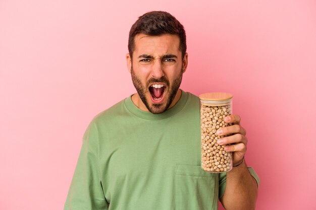 Young caucasian man holding a chickpea bottle isolated on pink background screaming very angry and aggressive.