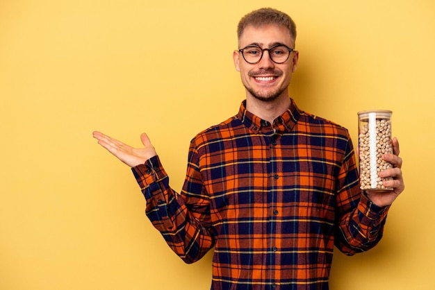 Young caucasian man holding chickpeas isolated on yellow background showing a copy space on a palm and holding another hand on waist.