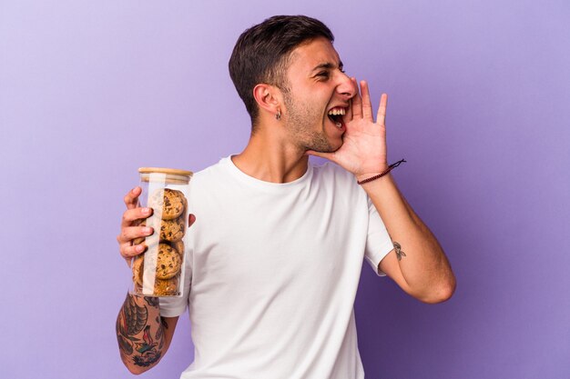 Молодой кавказский мужчина держит шоколадное печенье, изолированное на фиолетовом фоне, кричит и держит ладонь возле открытого рта.