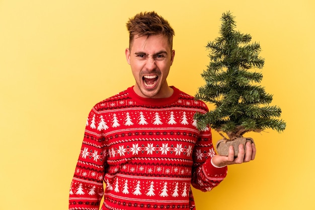 Young caucasian man holding a Christmas tree isolated on yellow background screaming very angry and aggressive.