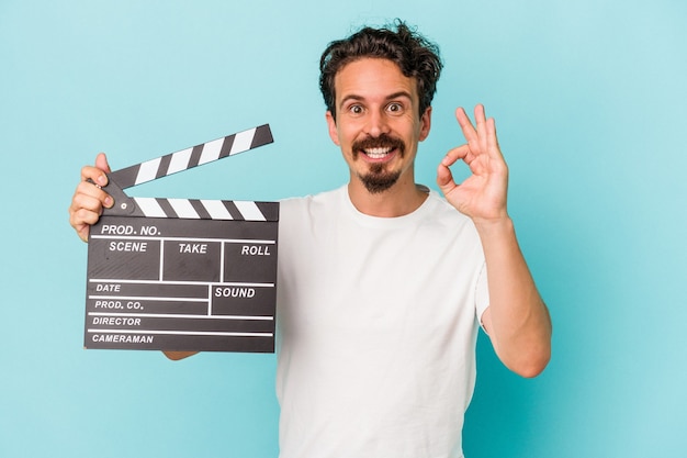 Young caucasian man holding clapperboard isolated on blue background cheerful and confident showing ok gesture.