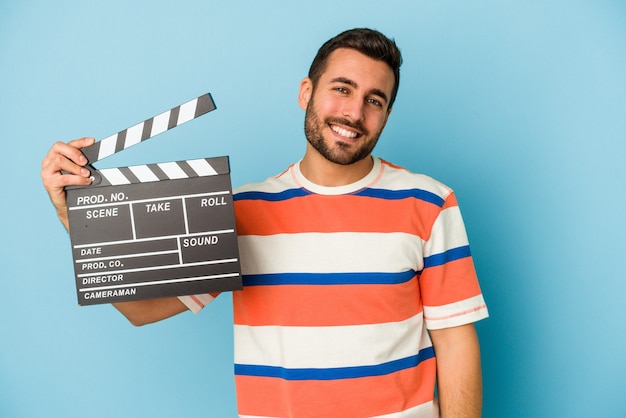 Foto giovane uomo caucasico che tiene un ciak isolato su sfondo blu felice, sorridente e allegro.