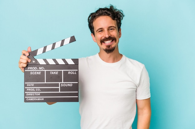 Young caucasian man holding clapperboard isolated on blue background happy, smiling and cheerful.