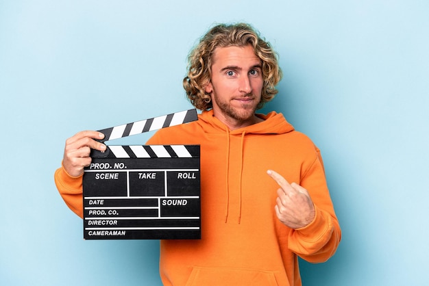 Young caucasian man holding a clapperboard isolated on blue background pointing with finger at you as if inviting come closer.