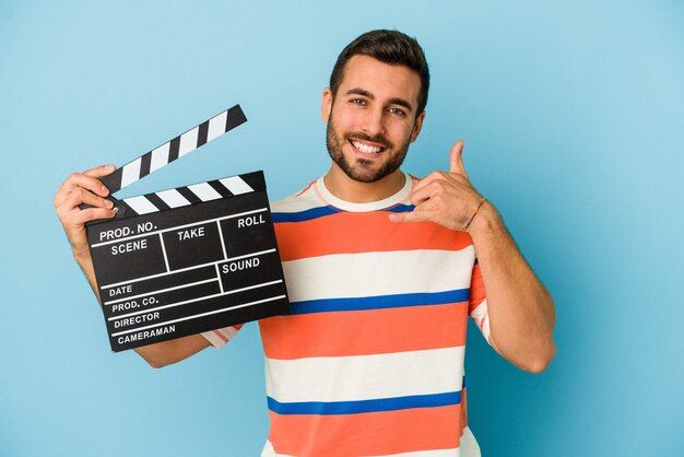 Young caucasian man holding a clapperboard isolated on blue background showing a mobile phone call gesture with fingers.