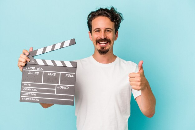 Young caucasian man holding clapperboard isolated on blue background smiling and raising thumb up