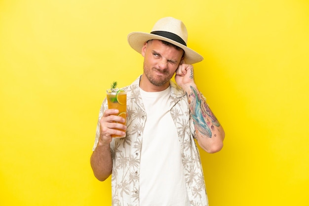 Young caucasian man holding a cocktail isolated on yellow background frustrated and covering ears