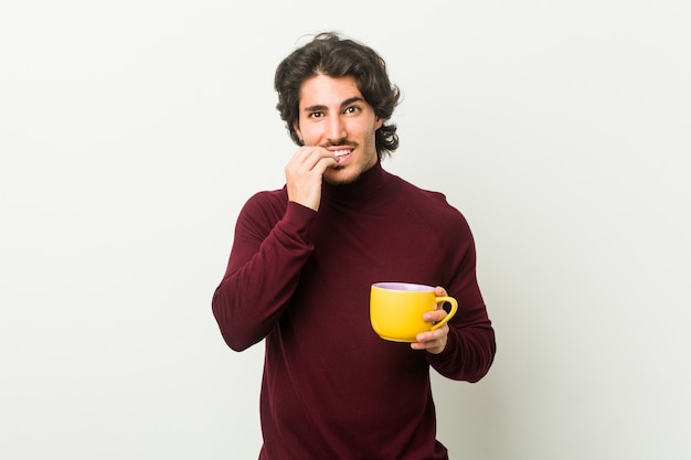 Young caucasian man holding a coffee cup biting fingernails, nervous and very anxious.