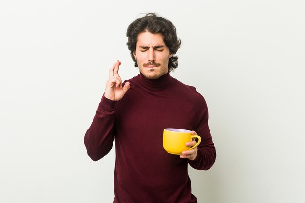 Young caucasian man holding a coffee cup crossing fingers for having luck