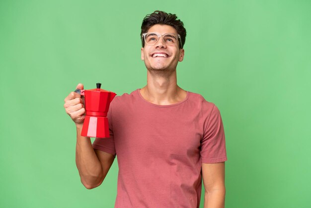 Young caucasian man holding coffee pot over isolated background looking up while smiling