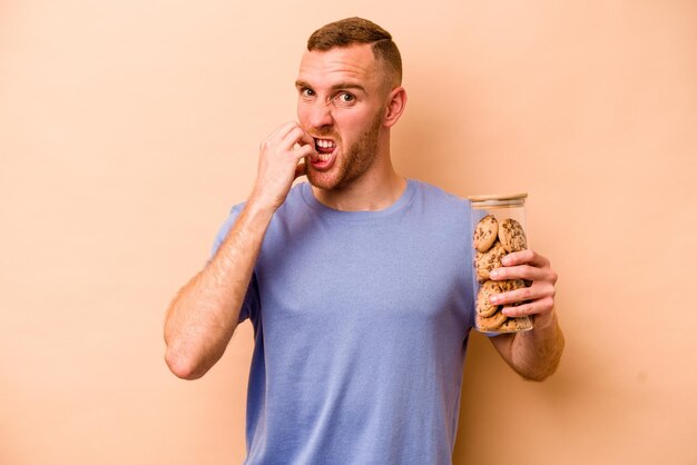 Young caucasian man holding cookies jar isolated on beige background biting fingernails nervous and very anxious