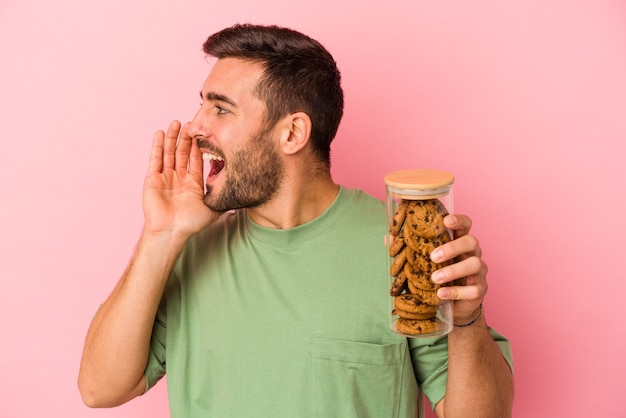 Young caucasian man holding cookies jar isolated on pink background shouting and holding palm near opened mouth.