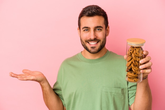 Young caucasian man holding cookies jar isolated on pink background showing a copy space on a palm and holding another hand on waist.