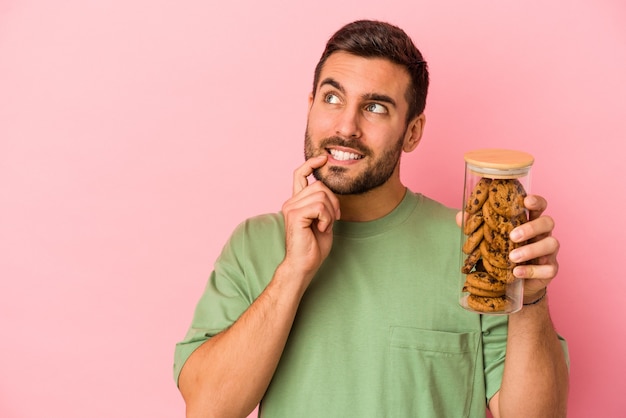 Young caucasian man holding cookies jar isolated on pink wall relaxed thinking about something looking at a copy space