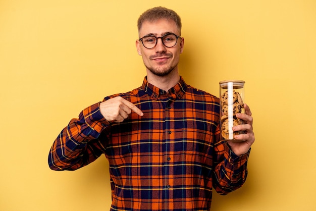 Young caucasian man holding cookies jar isolated on yellow background person pointing by hand to a shirt copy space, proud and confident