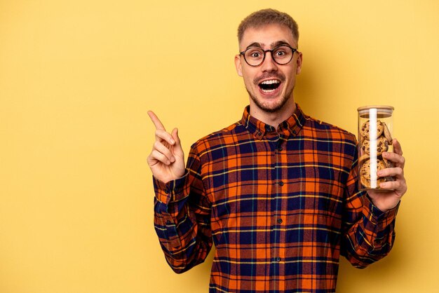 Young caucasian man holding cookies jar isolated on yellow background pointing to the side