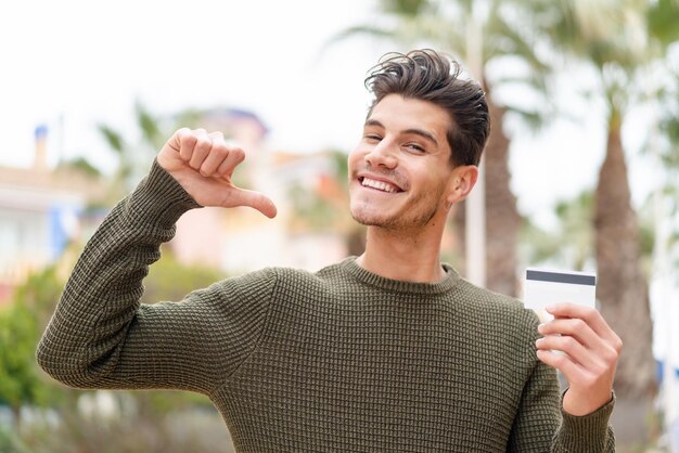 屋外でクレジットカードを持っている若い白人男性は、誇りに思って自己満足しています