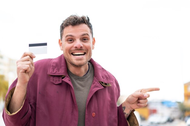 Young caucasian man holding a credit card at outdoors surprised and pointing finger to the side