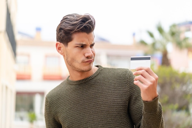 Young caucasian man holding a credit card at outdoors with sad expression